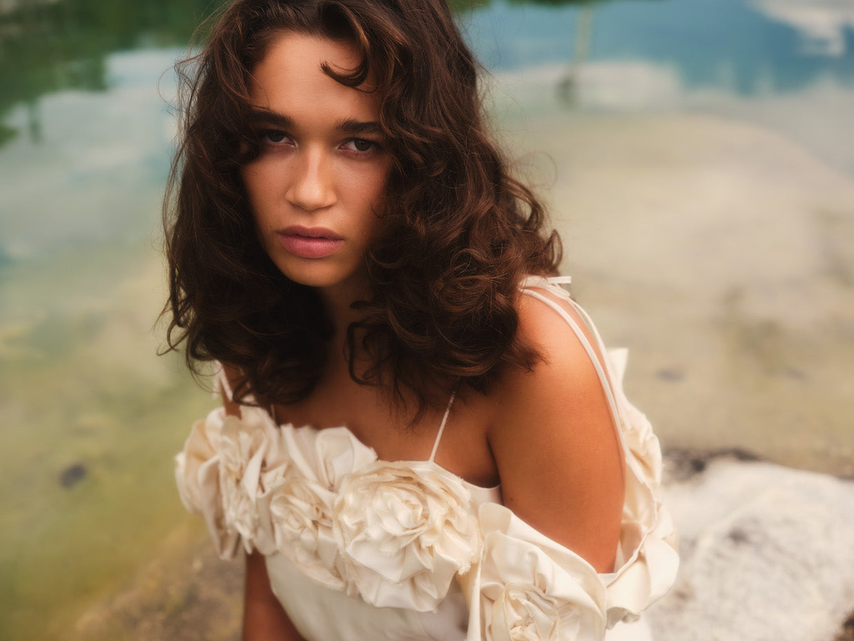 A woman with long, curly hair is wearing the Rosie Dress by Noémiah, which features a rose-embellished neckline. She is seated outdoors beside a calm body of water, gazing directly at the camera with a serene expression.