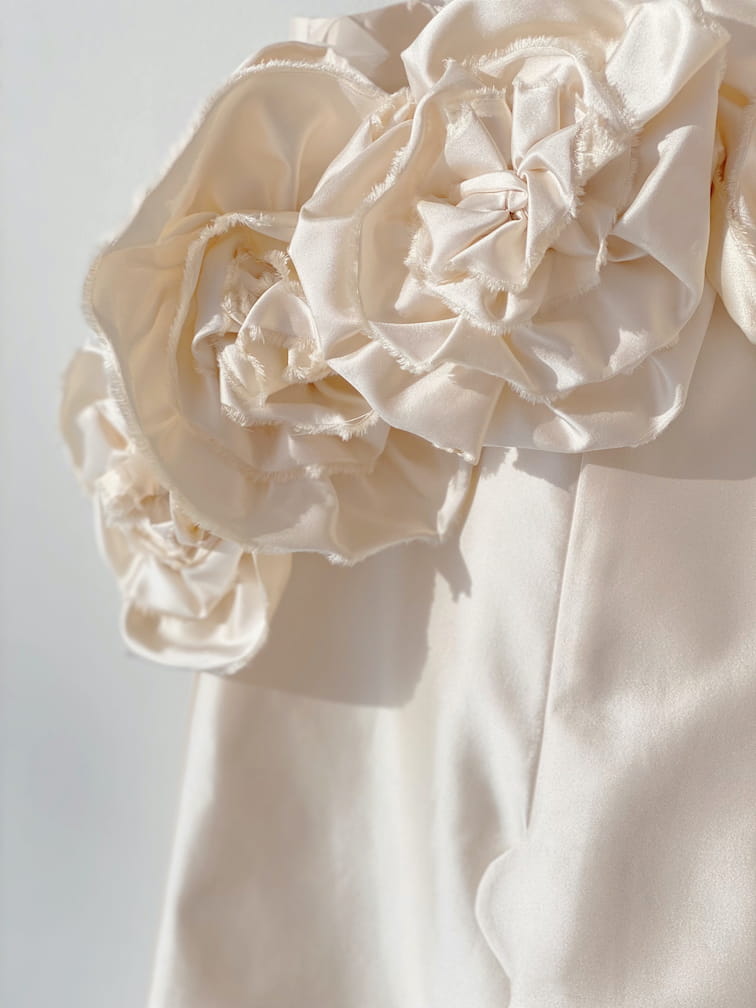 A close-up of the Rosie Dress by Noémiah shows a silk taffeta mini dress adorned with large, intricately crafted fabric flowers. The rose-embellished neckline adds a delicate and elegant touch to the dress, with petals that have a soft, textured appearance while the softly blurred background enhances the details.