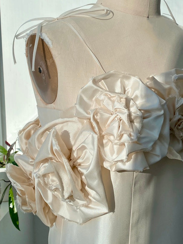 A close-up of the Rosie Dress by Noémiah on a mannequin showcases large, textured fabric flowers crafted from shiny, cream-colored silk taffeta. This mini dress features slim shoulder straps and is ideal for a wedding gown. It is displayed in natural light, casting gentle shadows over its rose-embellished neckline.