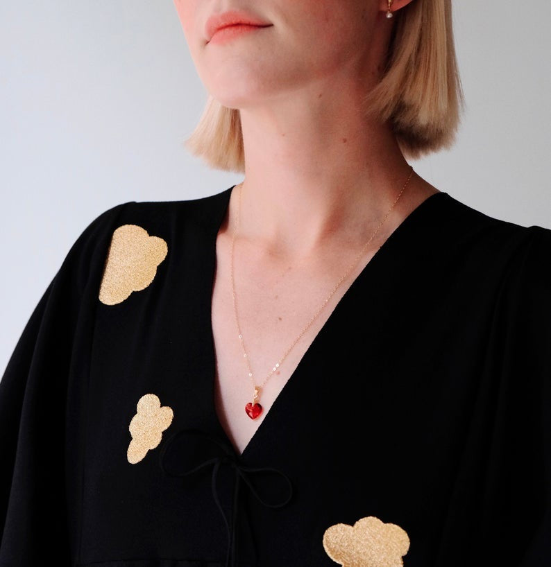 A person with short blond hair wears a black top adorned with gold cloud patterns. A Noémiah Rosita Pendant, featuring a sparkling red heart shape, graces their delicate gold-filled chain. The background is a clean, plain white.