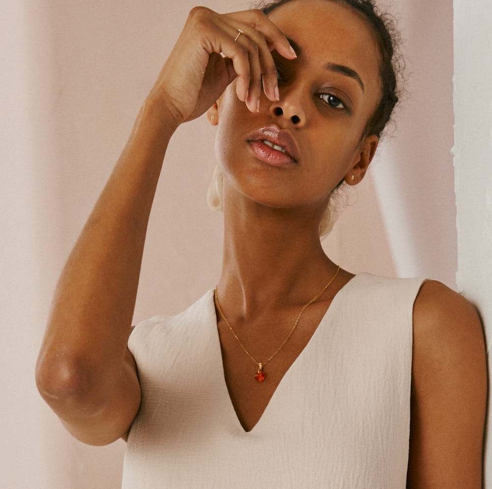 A woman with her hand partially covering her face stands against a light pink background, wearing a sleeveless beige top and the Rosita Pendant by Noémiah, featuring a gold-filled chain with a small diamond.