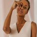 A woman with her hand partially covering her face stands against a light pink background, wearing a sleeveless beige top and the Rosita Pendant by Noémiah, featuring a gold-filled chain with a small diamond.