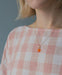 A woman wearing the Noémiah Rosita Pendant, featuring a gold-filled chain and a delicate orange Swarovski crystal. She is dressed in a pink and white checkered top set against a simple grayish-blue background.