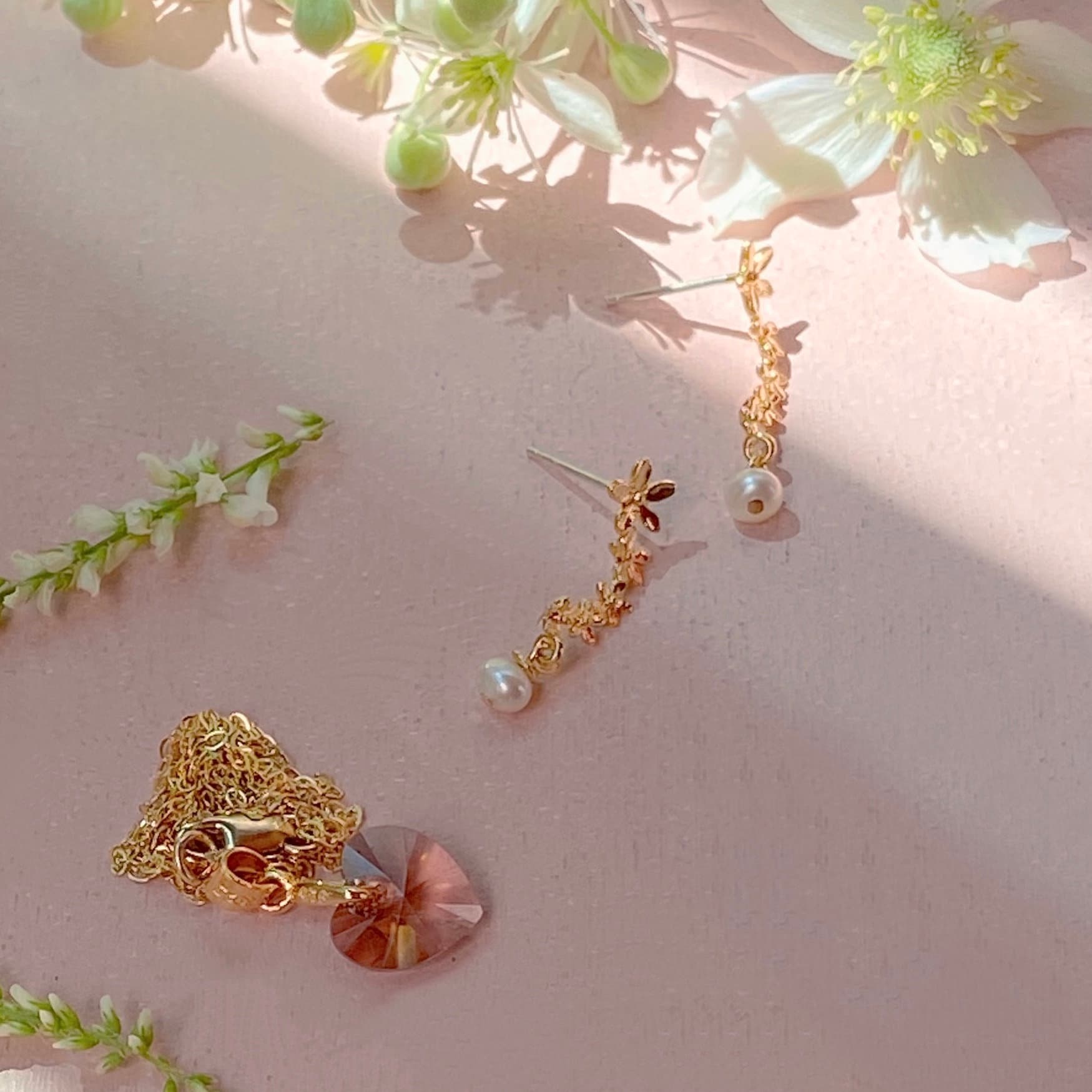 The Rosita Pendant by Noémiah, featuring a heart-shaped Swarovski crystal, is displayed alongside delicate gold earrings adorned with pearl accents. These elegant pieces rest on a pink surface, surrounded by softly focused flowers that enhance the light and airy ambiance.