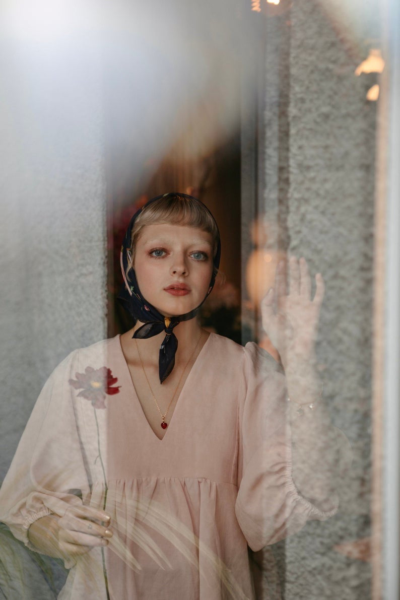 A woman in a light pink dress and headscarf stands behind a glass window, delicately holding a flower. She gazes thoughtfully outside as her hand, adorned with the Rosita Pendant by Noémiah, touches the glass. Reflections and soft, diffused lighting create a serene atmosphere.