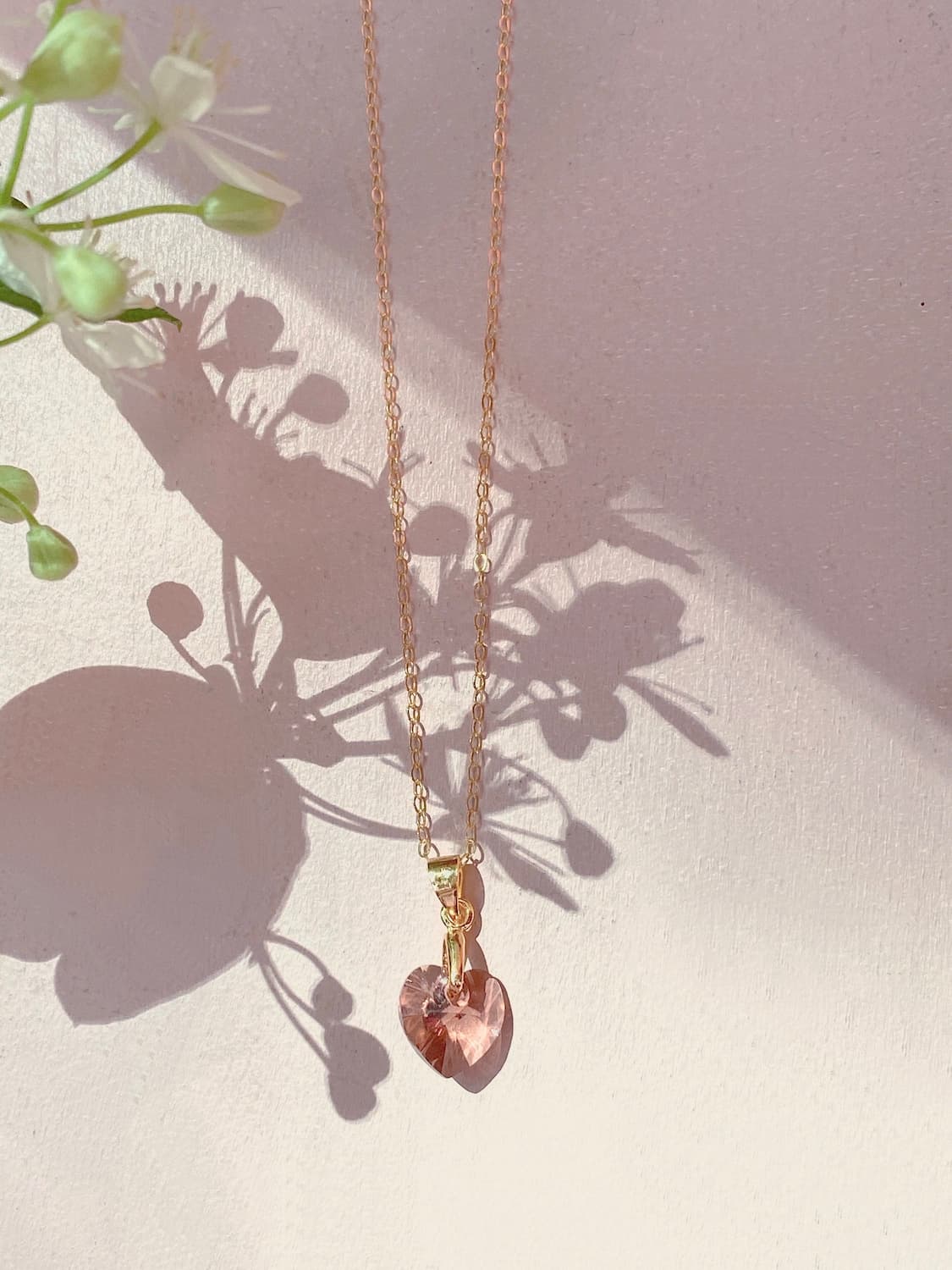 The Rosita Pendant by Noémiah, featuring a gold-filled chain and a heart-shaped pink Swarovski crystal, hangs elegantly against a light surface. Intricate patterns are formed by the shadow of the necklace and the surrounding greenery.