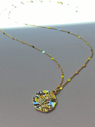 The Sabrina Pendant from Noémiah, a gold-plated necklace with a delicate chain accented by small colored beads, is displayed on a flat surface. The pendant resembles a vintage coin charm and is adorned with an intricate floral design depicting a rose.