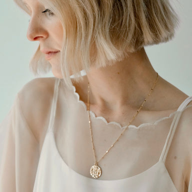 A woman with short blonde hair looks down, wearing a sheer white top with scalloped edges. She adorns the Sabrina Pendant by Noémiah, a gold-plated necklace featuring a vintage coin charm with a leaf design. The background is soft and neutral.