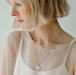 A woman with short blonde hair looks down, wearing a sheer white top with scalloped edges. She adorns the Sabrina Pendant by Noémiah, a gold-plated necklace featuring a vintage coin charm with a leaf design. The background is soft and neutral.