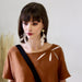 A woman with long brown hair is wearing a brown top, embellished with high-quality feathers on the shoulders, and large Salomé Earrings from Noémiah that are tarnish-resistant. She has a black strap across her chest from a bag and stands against a light background.