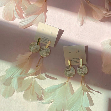 Two pairs of Salomé Earrings by Noémiah, featuring circular studs and long, delicate white feathers, are displayed on a soft pink surface. These lightweight earrings boast tarnish-resistant plating and are attached to white cards adorned with the n.h brand logo, surrounded by additional loose feathers.