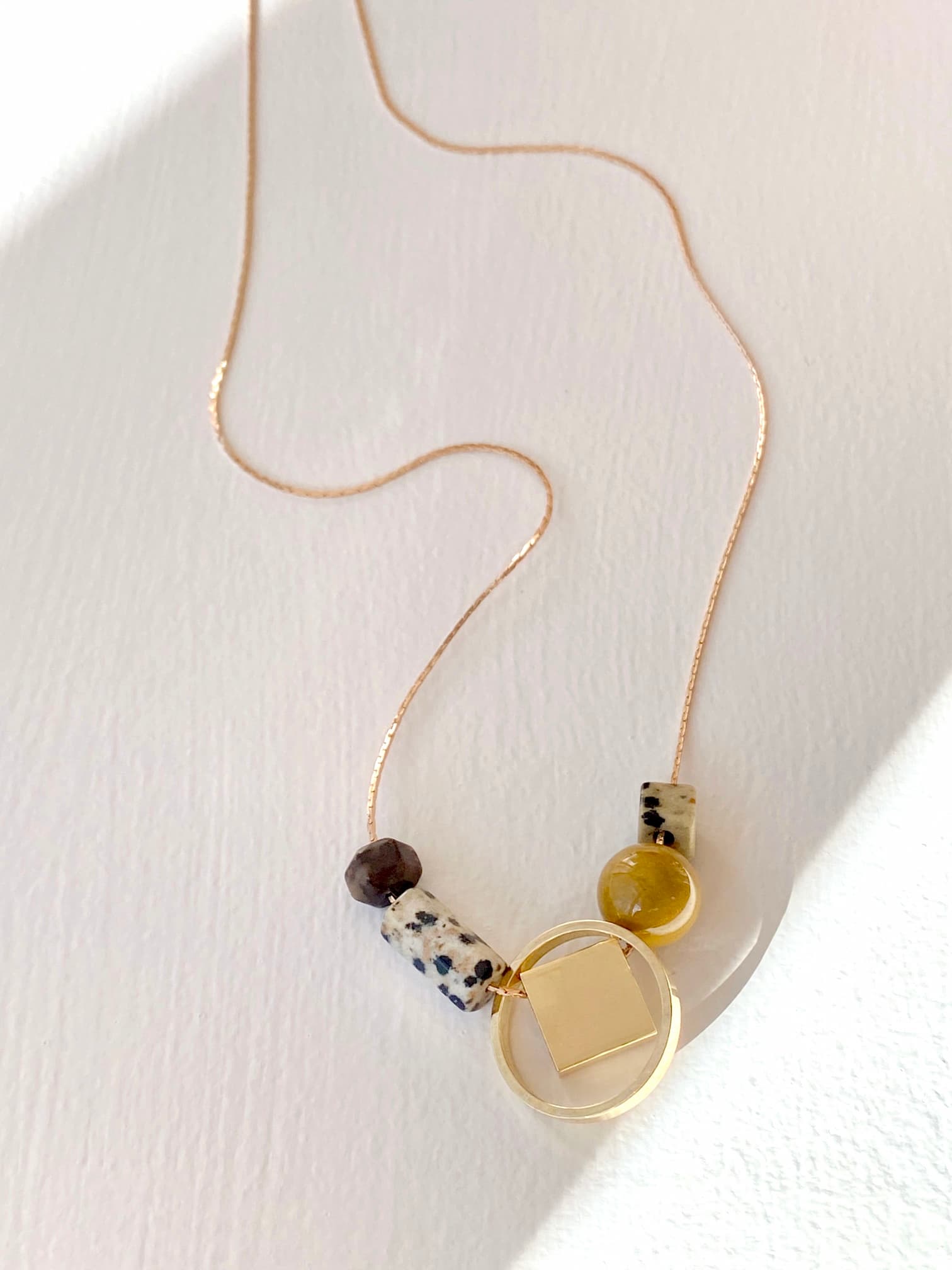 The Samuel Necklace by Noémiah is a delicate piece featuring a thin gold chain and minimalist design, adorned with unique beads such as a brown bead, a yellow circular bead, and a tarnish-resistant gold geometric pendant. It rests on the white surface, where the lighting casts soft shadows.