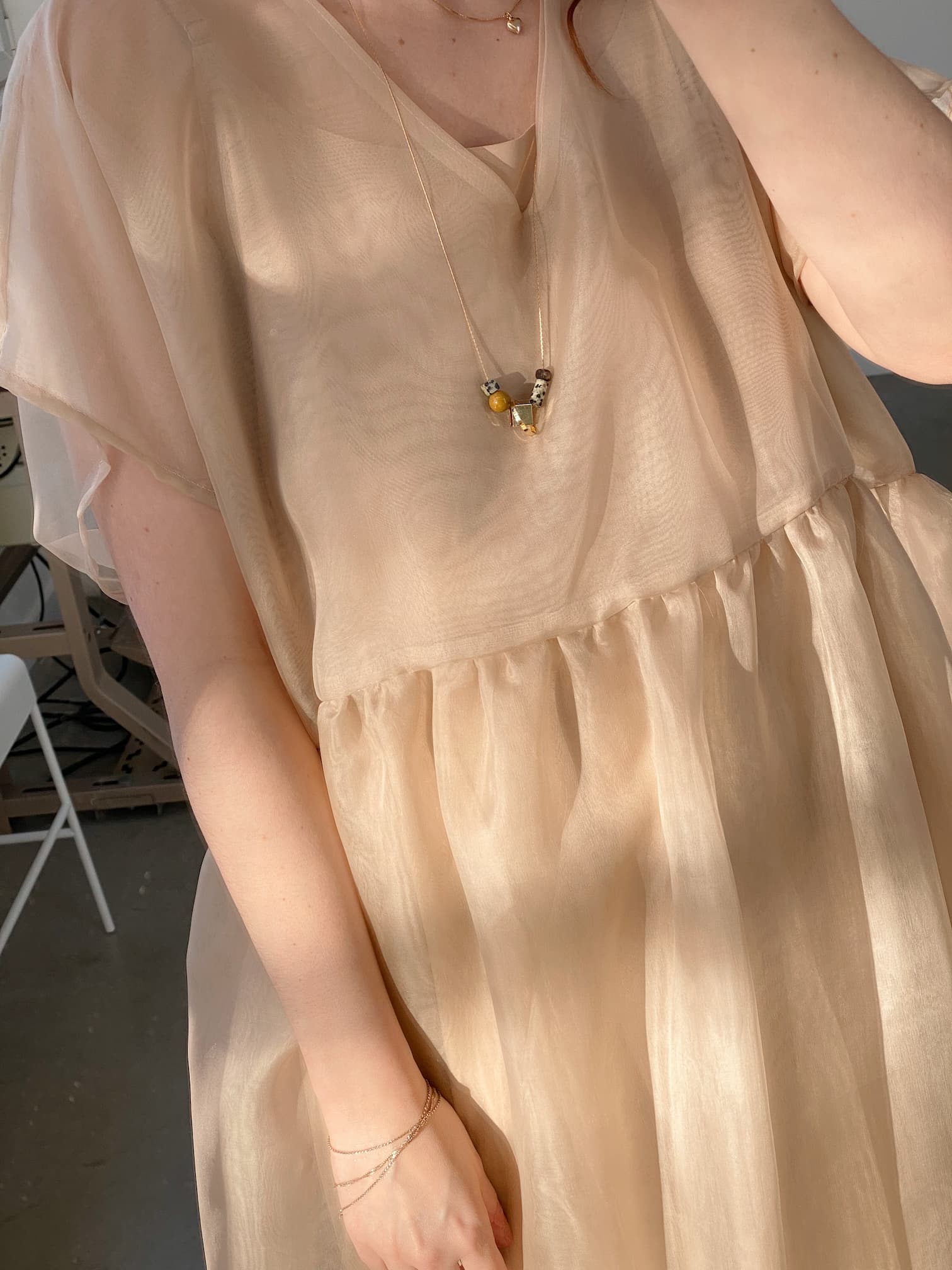 A person wearing a flowy, semi-transparent beige dress is adorned with the Samuel Necklace by Noémiah, which features small gemstone details. Their arm is partially visible as sunlight casts soft shadows across the fabric, and in the background stands a tarnish-resistant white chair.