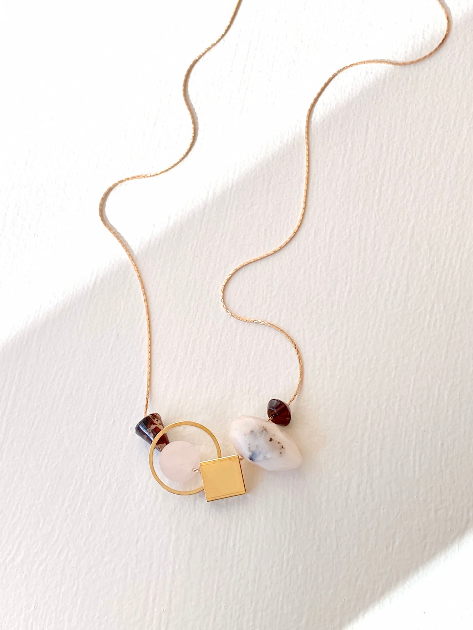 The Samuel Necklace by Noémiah is a delicate gold piece featuring a geometric pendant adorned with a gold square, circle, and marbled stones. This minimalist design is tarnish-resistant and elegantly displayed on a light background with soft shadows.