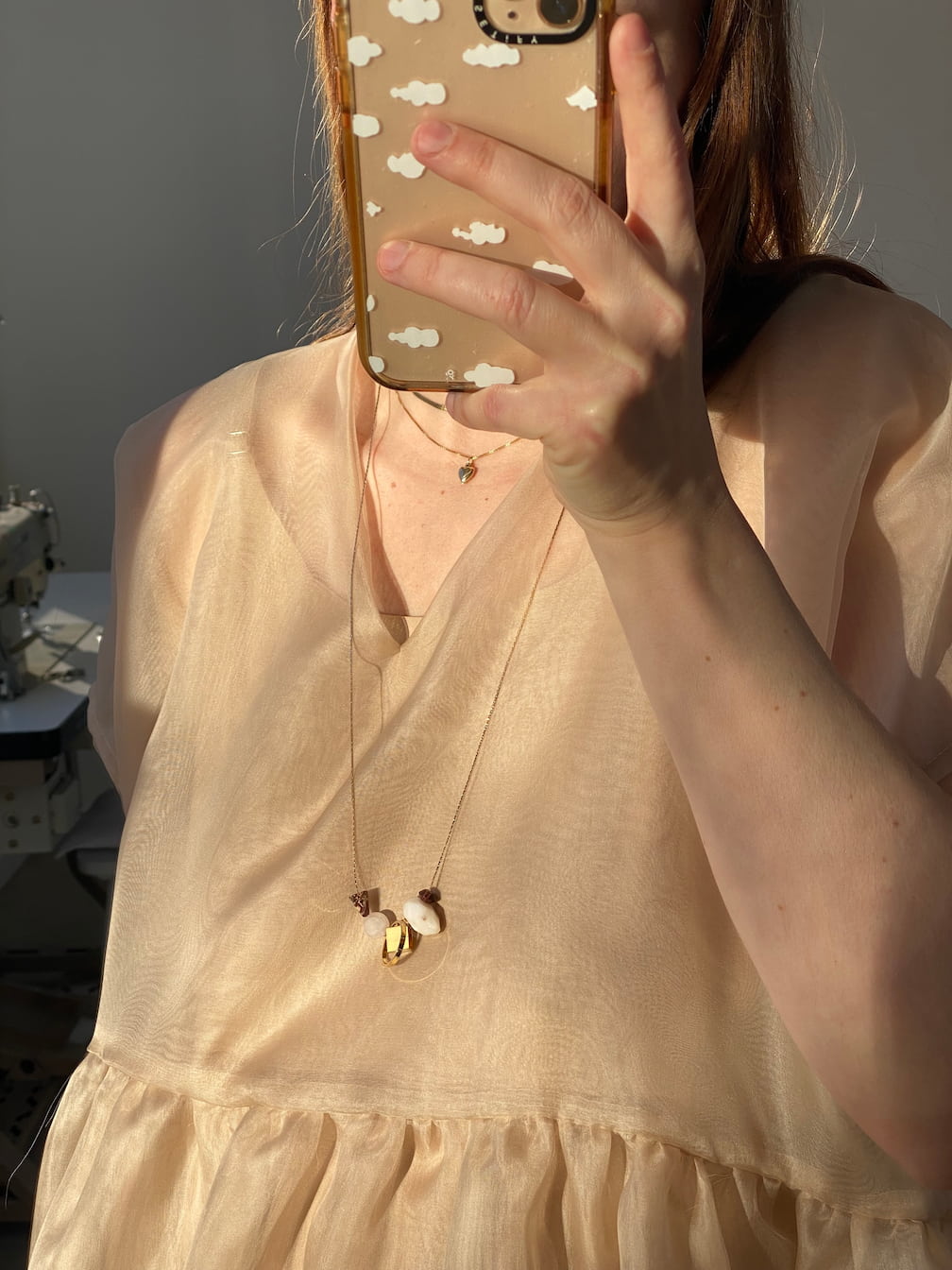 A person takes a mirror selfie wearing a sheer, light beige blouse. The smartphone case features white cloud patterns. They accessorize with the tarnish-resistant Samuel Necklace by Noémiah, which is adorned with gemstone details. Sunlight casts soft shadows on their outfit.