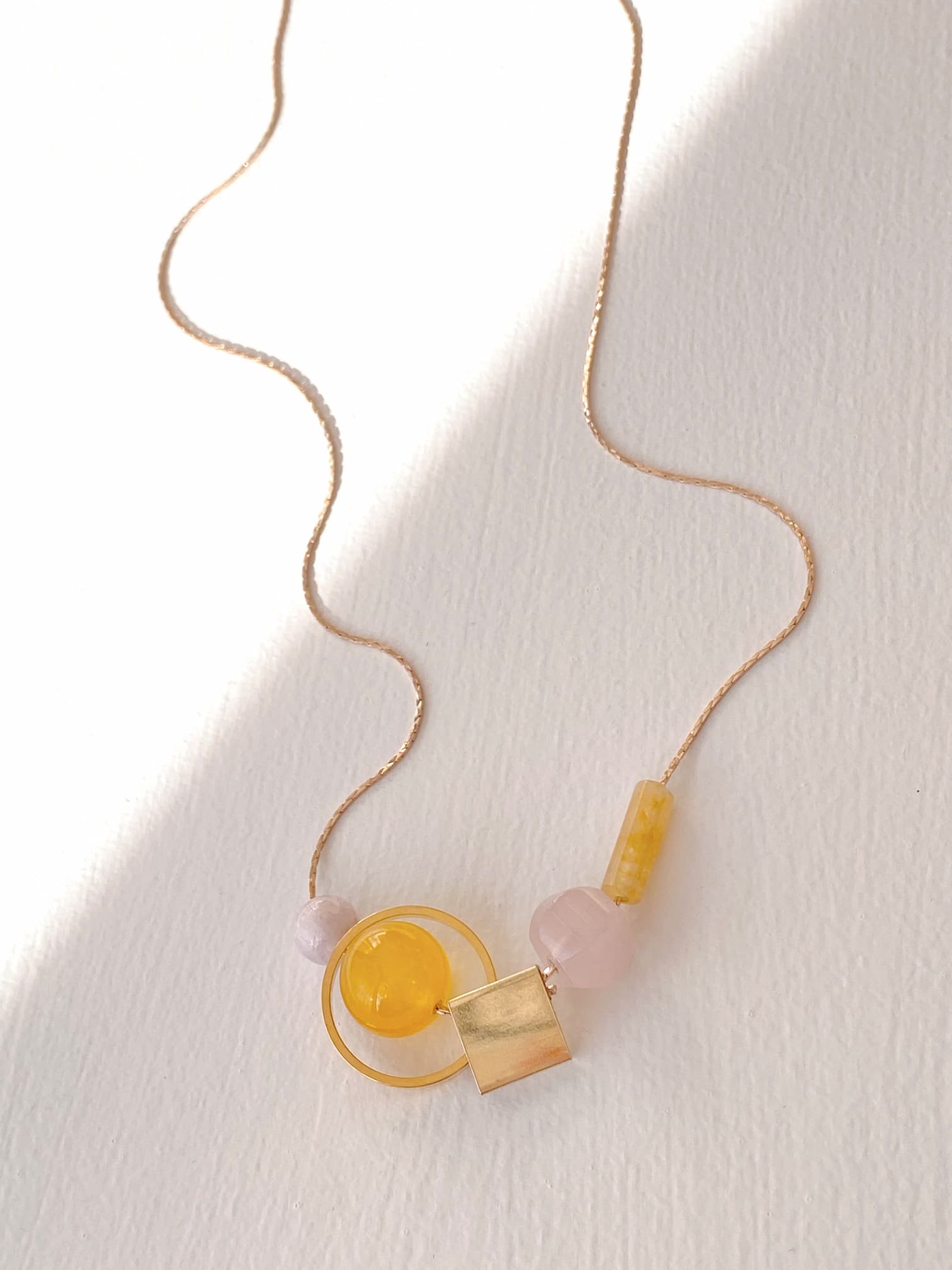 The Samuel Necklace by Noémiah features a delicate and tarnish-resistant thin gold chain, embodying minimalist design with its artful arrangement of several beads: a translucent yellow sphere, a yellow rectangular bead, and a gold square. Displayed elegantly on a light surface, it casts soft shadows that enhance its sophisticated charm.