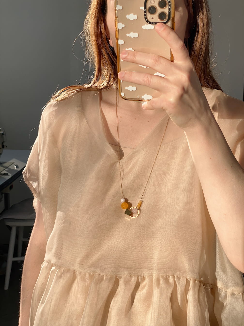 Dressed in a sheer, light beige blouse, a person takes a mirror selfie with their phone adorned by a case featuring cloud designs. Around their neck hangs the Samuel Necklace by Noémiah, a tarnish-resistant piece with minimalist design and gemstone details including an orange bead.