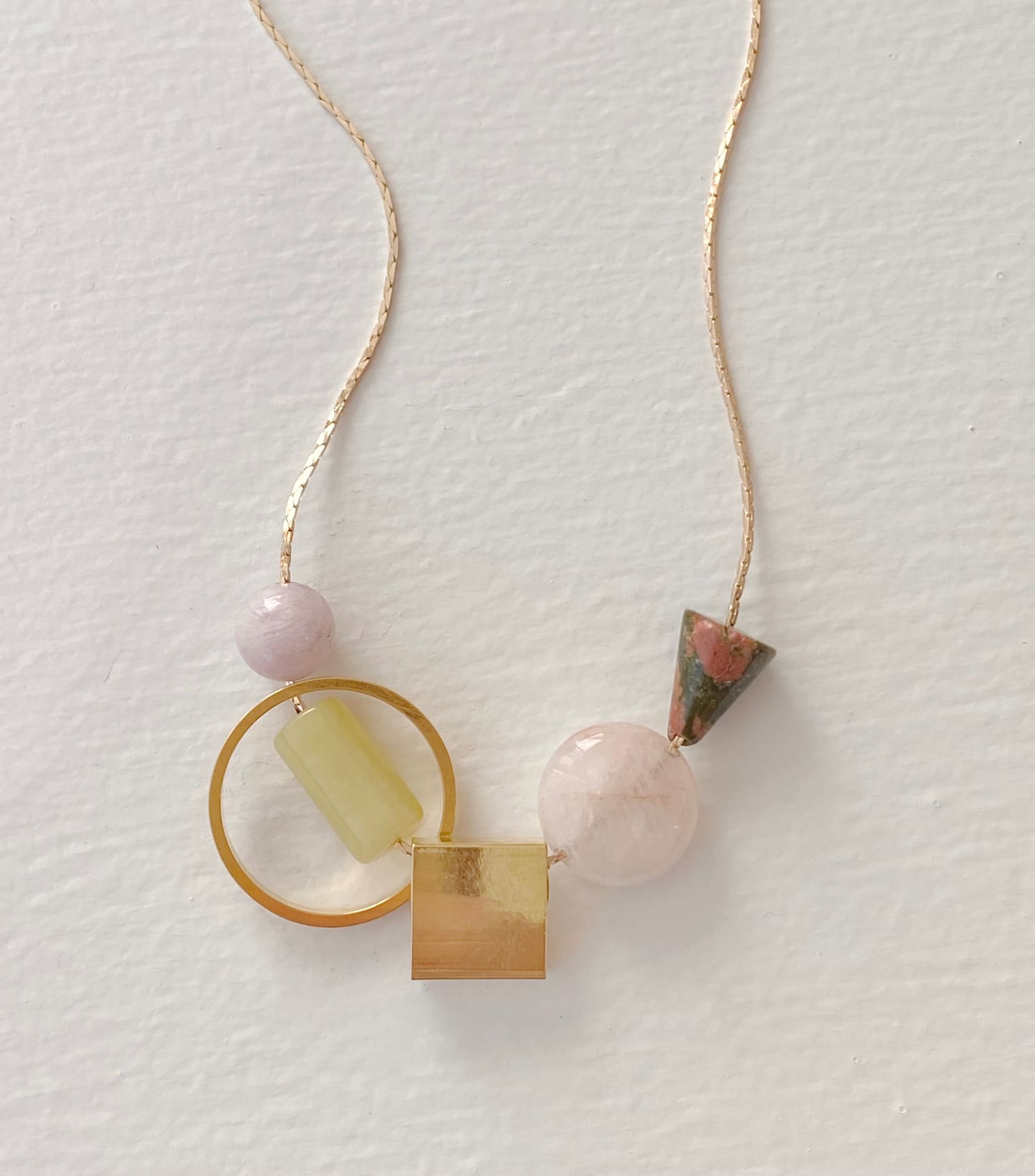 The Samuel Necklace by Noémiah is an elegant piece with a tarnish-resistant gold chain, adorned with an array of geometric beads including a yellow cylinder, a pink sphere, a green cone, a square gold piece, and a gold circle. Its playful and minimalist design effortlessly enhances any outfit.