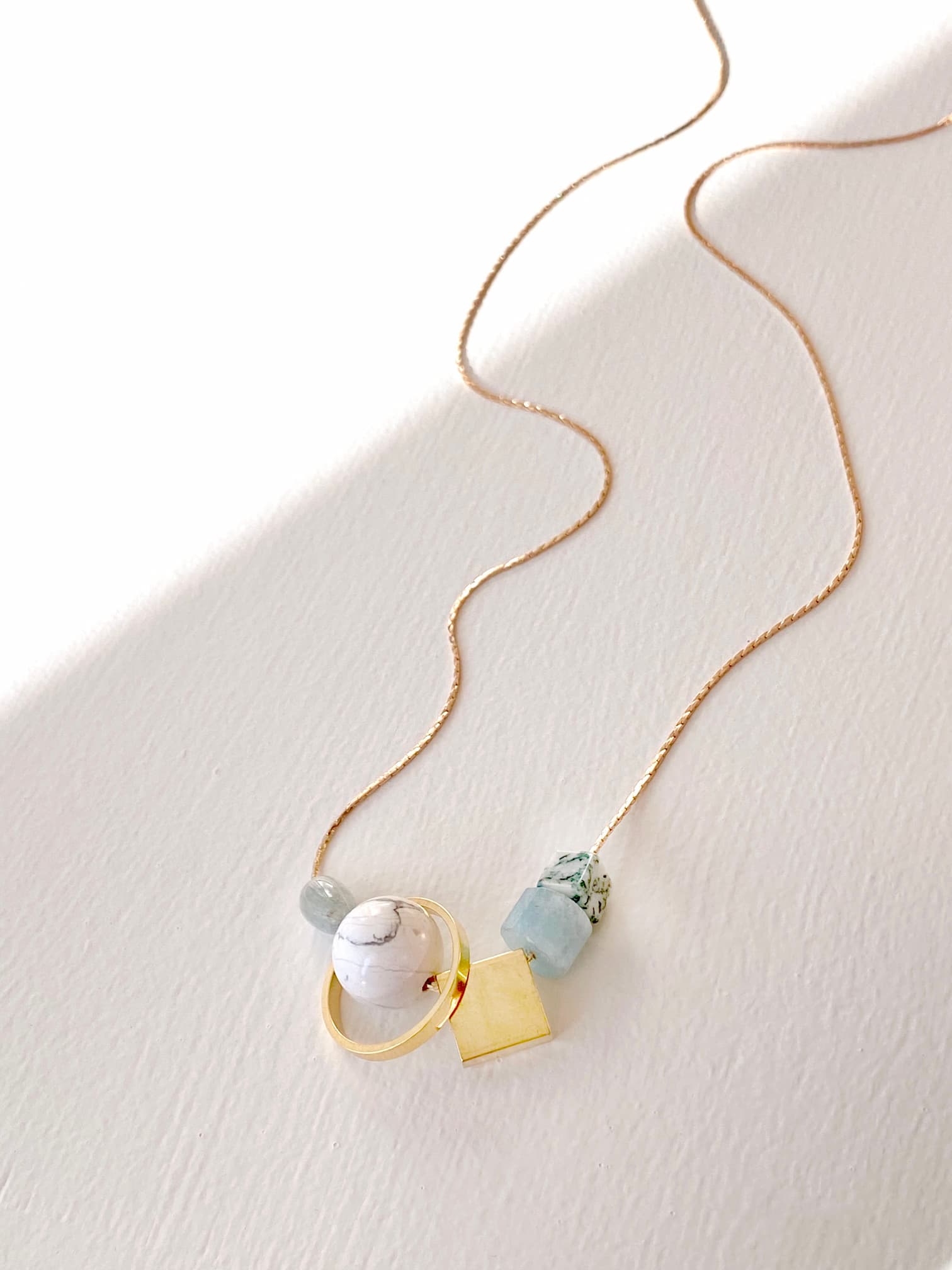 The Samuel Necklace by Noémiah features a delicate, tarnish-resistant gold chain adorned with a pendant consisting of a round white stone, a small green gemstone, and a light blue stone. Gold geometric details complete its minimalistic design, allowing this elegant piece to rest gracefully on a white surface.