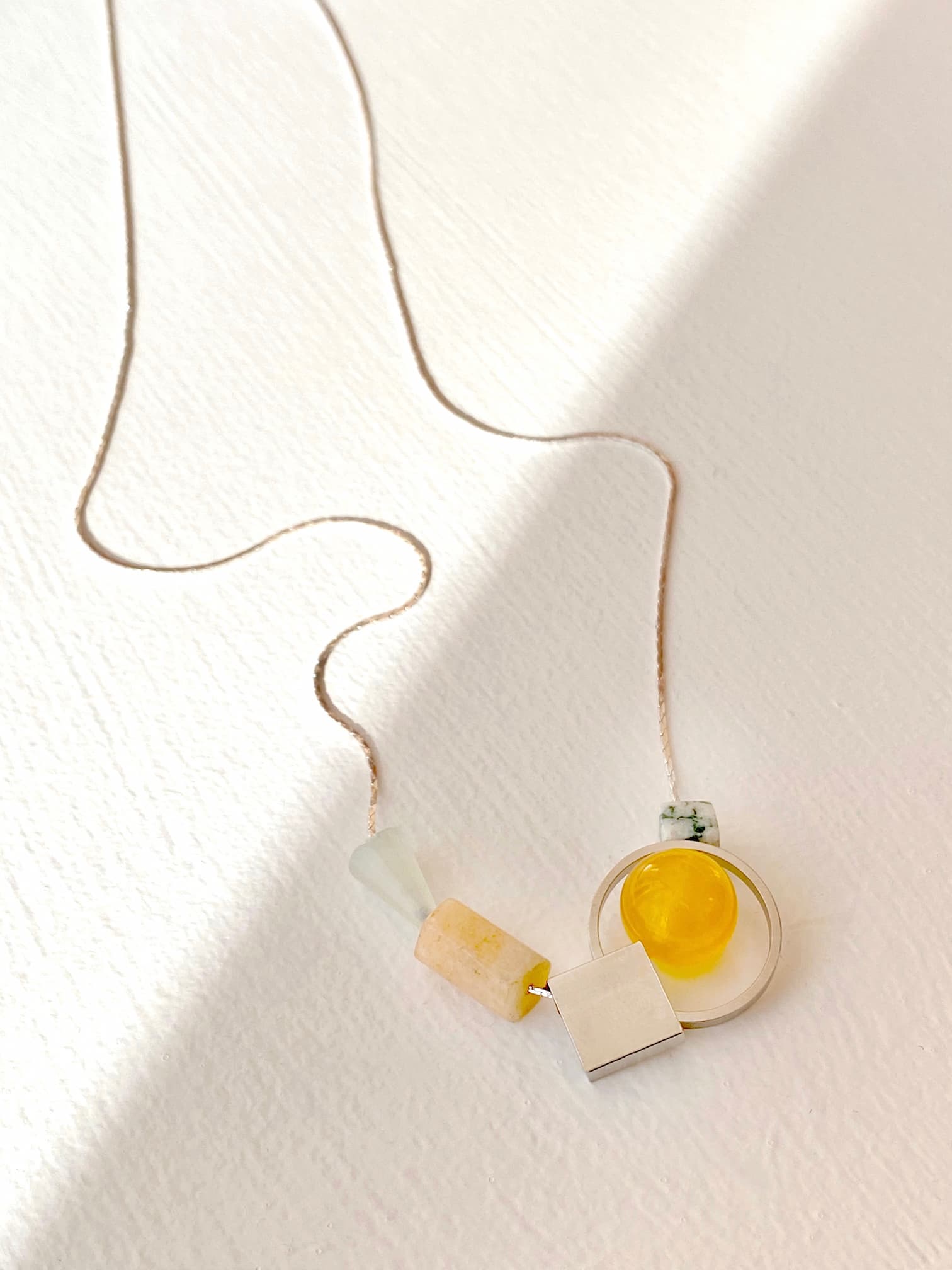 Explore the Samuel Necklace by Noémiah, featuring a tarnish-resistant design that captures minimalist elegance. This necklace showcases a slim chain adorned with geometric shapes, including a vibrant yellow circular piece and cream and white beads. Photographed on a light surface, it casts a gentle shadow for an added touch of sophistication.