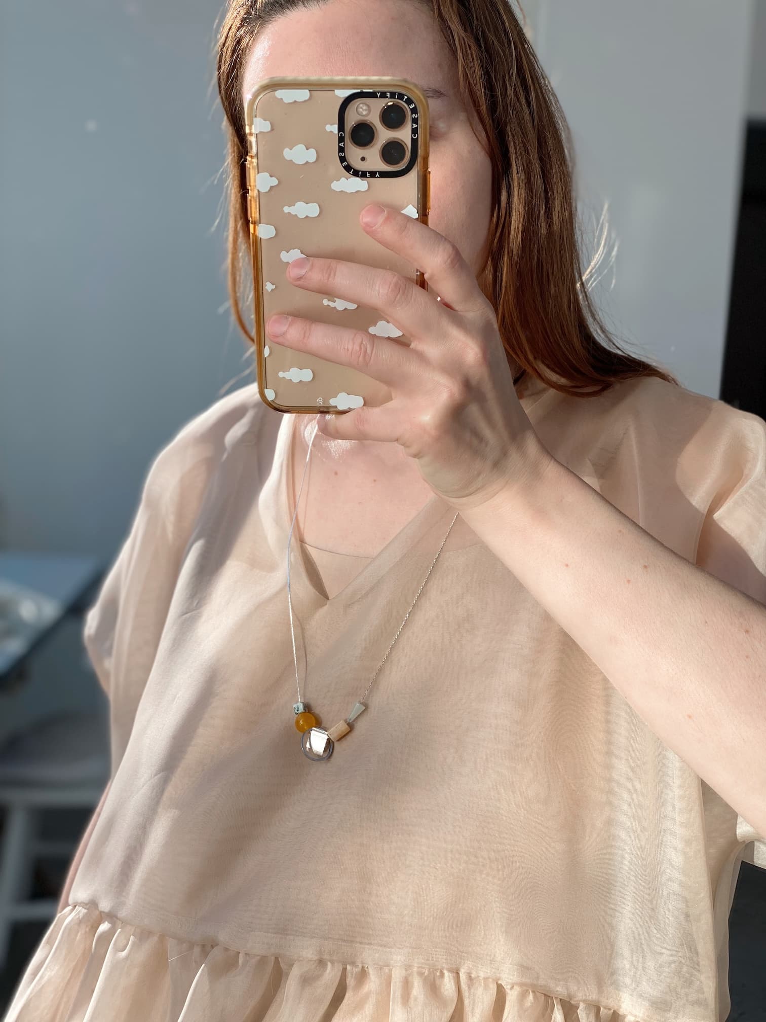 A person with long brown hair captures a mirror selfie using a phone encased in a cloud-patterned cover. They are dressed in a beige, loose-fitting blouse and adorned with the Samuel Necklace from Noémiah, which is tarnish-resistant and features minimalist gemstone details. The background appears softly blurred.