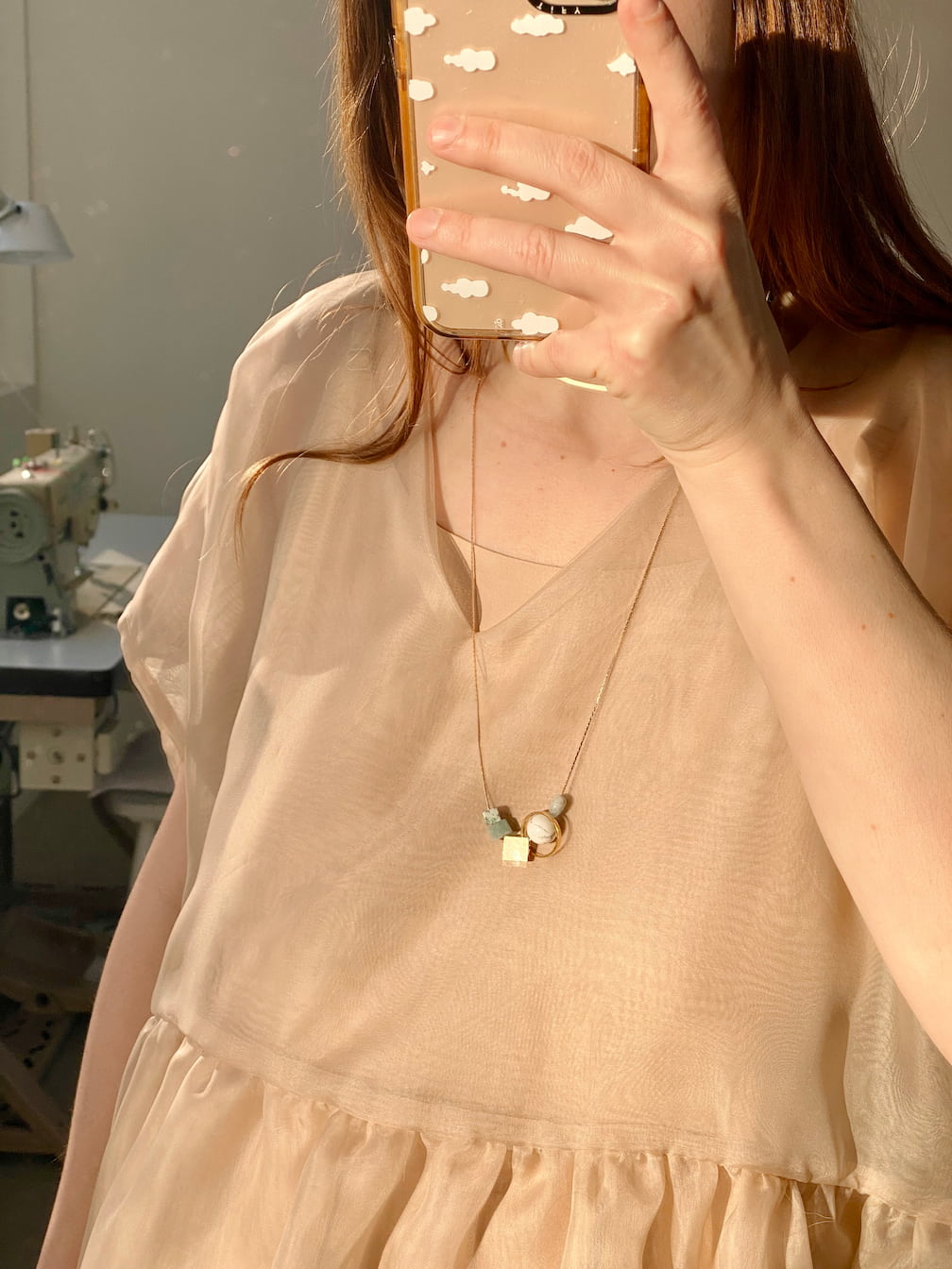 The photo features an individual taking a mirror selfie, dressed in a light, flowy beige dress and wearing the Samuel Necklace by Noémiah, which is embellished with gemstone details. The phone case boasts a cloud pattern. In the background, there's a sewing machine that complements the minimalist aesthetic of the image.