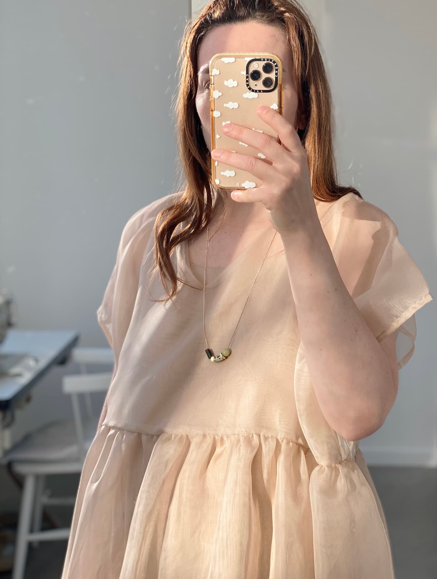 A person wearing a light, sheer dress is taking a mirror selfie indoors with a phone case featuring a cloud pattern. Sunlight softly illuminates the scene as they wear the Samuel Necklace by Noémiah, adorned with elegant gemstone details.