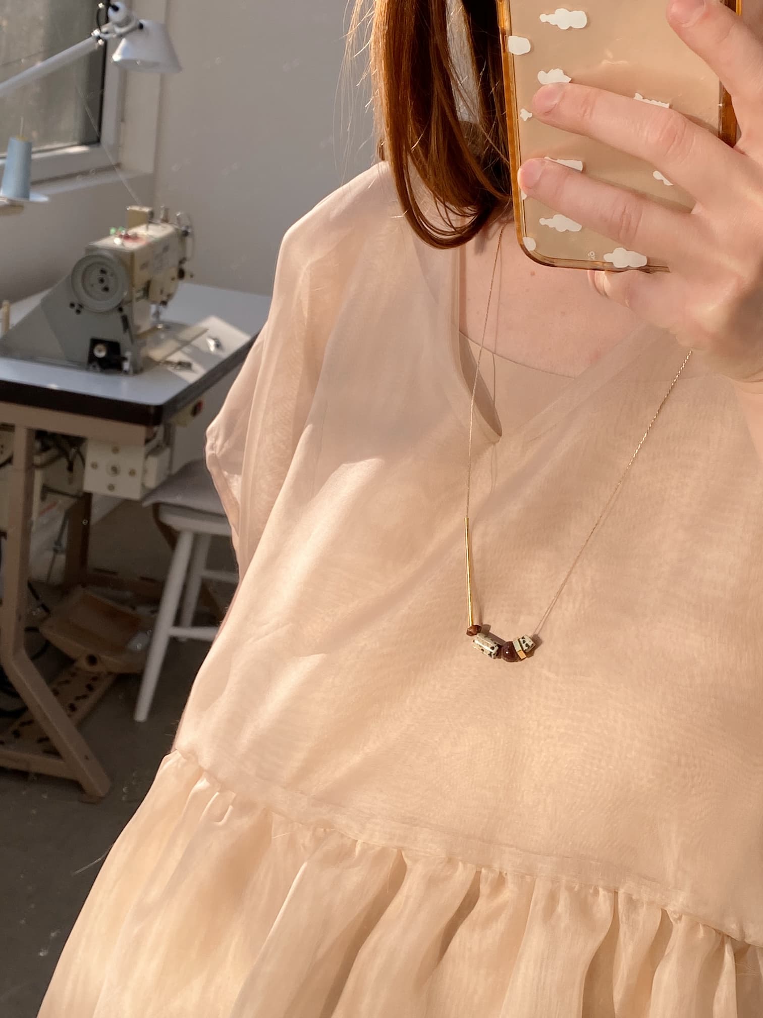 A person wearing a light, sheer dress takes a mirror selfie, holding a phone with a decorative case featuring gemstone details. Around their neck hangs the Samuel Necklace from Noémiah, known for its tarnish-resistant gold finish and bead adornments. In the background, a sewing machine can be seen near a window.