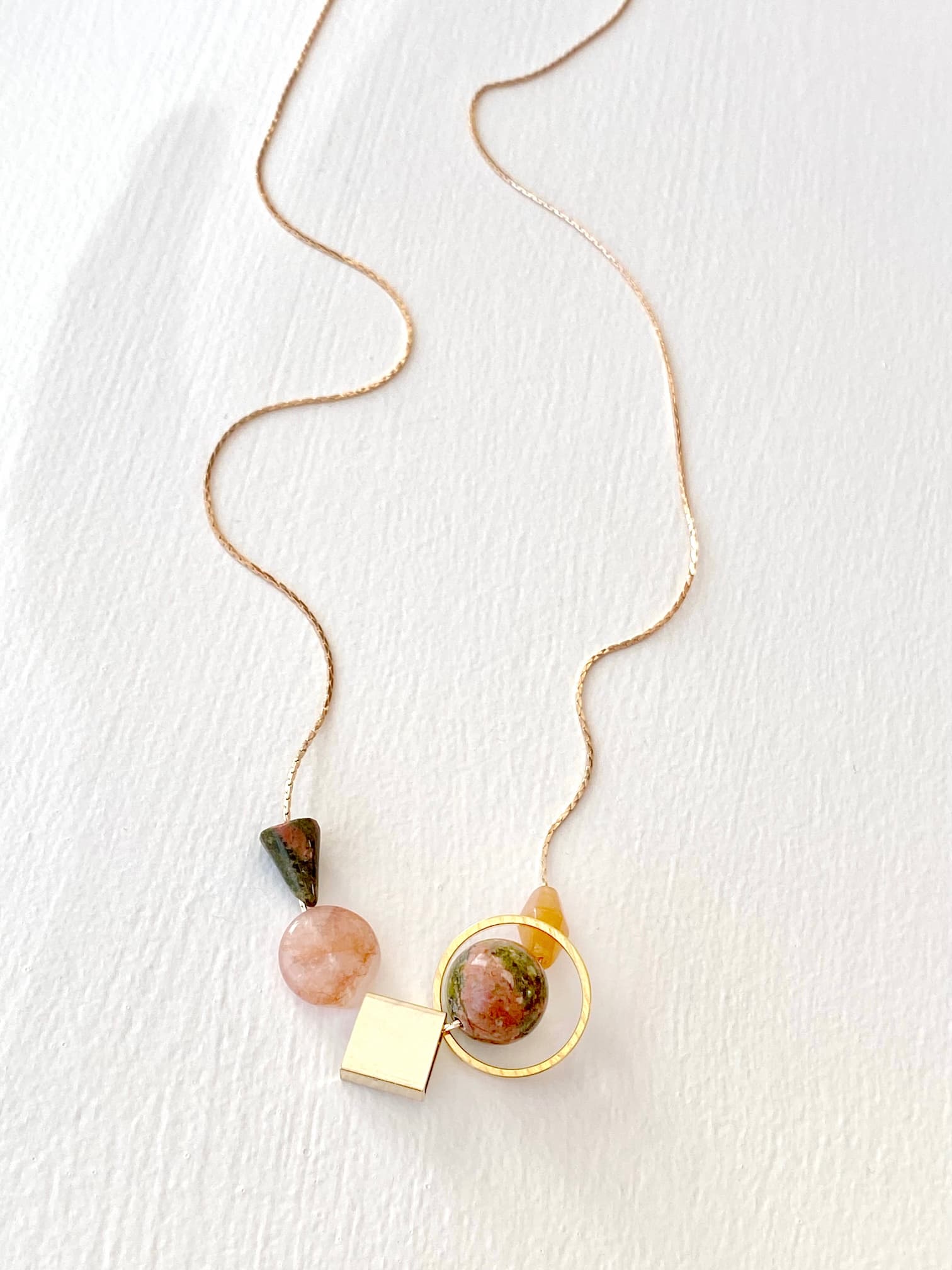 The Samuel Necklace by Noémiah is a delicate gold chain necklace with tarnish-resistant, minimalist design, featuring a selection of pendants: a pink circular stone, a small green triangular stone, a larger red and green circular stone in a gold ring, a square gold piece, and a tiny orange gem.