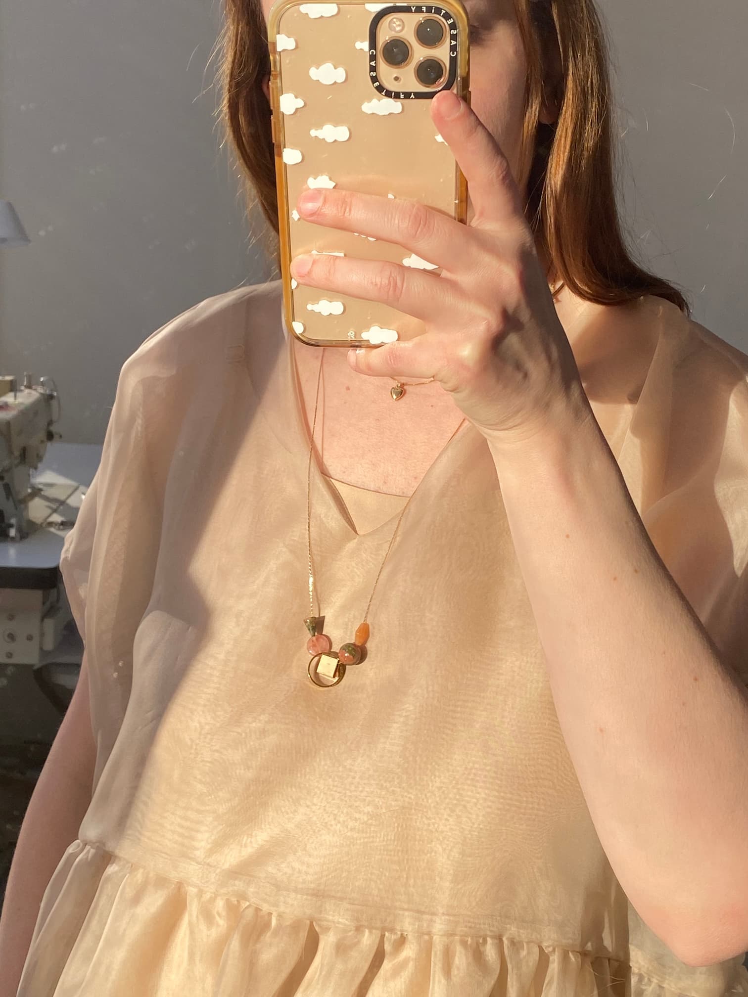 A person wearing a sheer, light-colored dress takes a mirror selfie. Their phone case features a white cloud pattern. They are also wearing the Samuel Necklace by Noémiah, which is a minimalist piece with a circular pendant and gemstone details, crafted to be tarnish-resistant for lasting elegance.