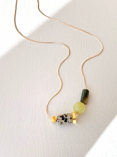 The Samuel Necklace by Noémiah features a minimalist design with three beads: a spotted stone, a round yellow bead, and a green cylindrical bead. This tarnish-resistant gold necklace is artistically arranged on a white background.