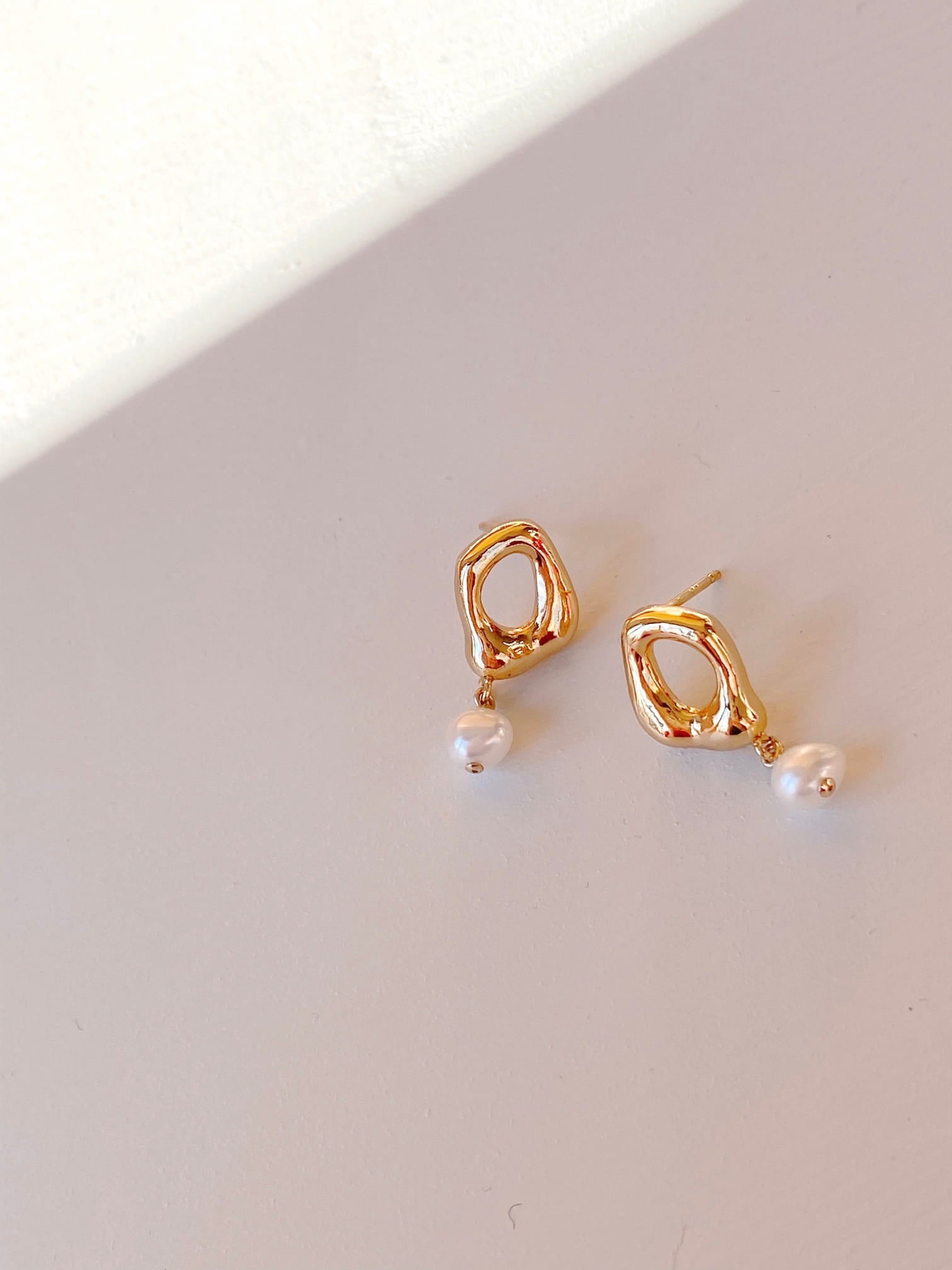 The Scarlett Earrings by Noémiah are a pair of gold hoops with organic shapes and irregular geometric designs, each featuring a dangling freshwater natural pearl, all placed on a light-colored surface with soft shadows.