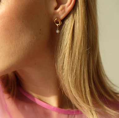 A person with light hair wears a pink top, showcasing the Scarlett Earrings from Noémiah—gold organic-shaped hoops adorned with a hanging freshwater natural pearl. Soft lighting highlights their shimmering earring and delicate neck area.