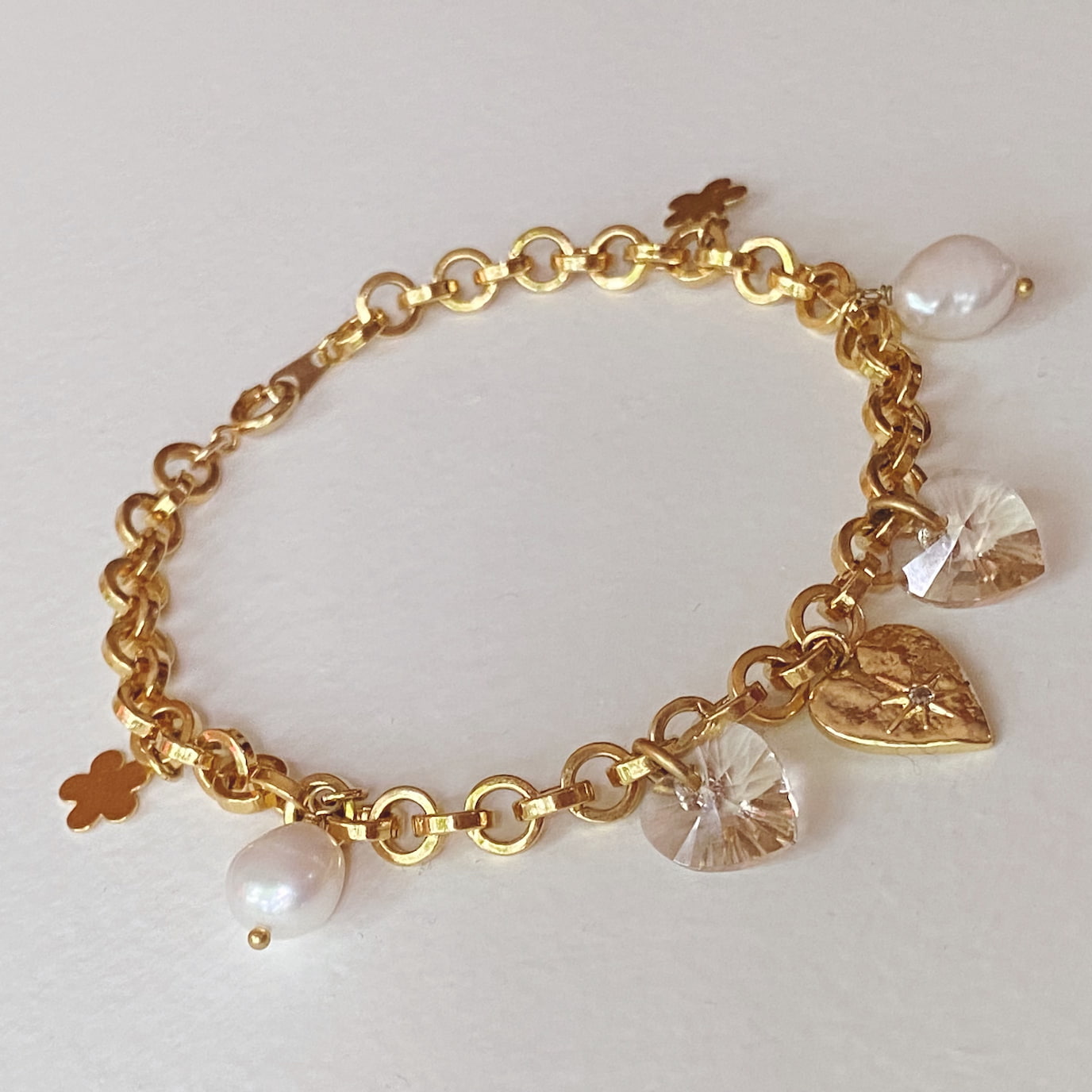 Presenting the Secret Heart Bracelet by Noémiah, a beautiful gold chain decorated with charms such as pearls and floral designs. The heart charm is enhanced with sparkling Swarovski crystals and intricate patterns, exuding timeless elegance on a white background. This tarnish-resistant piece is truly stunning.