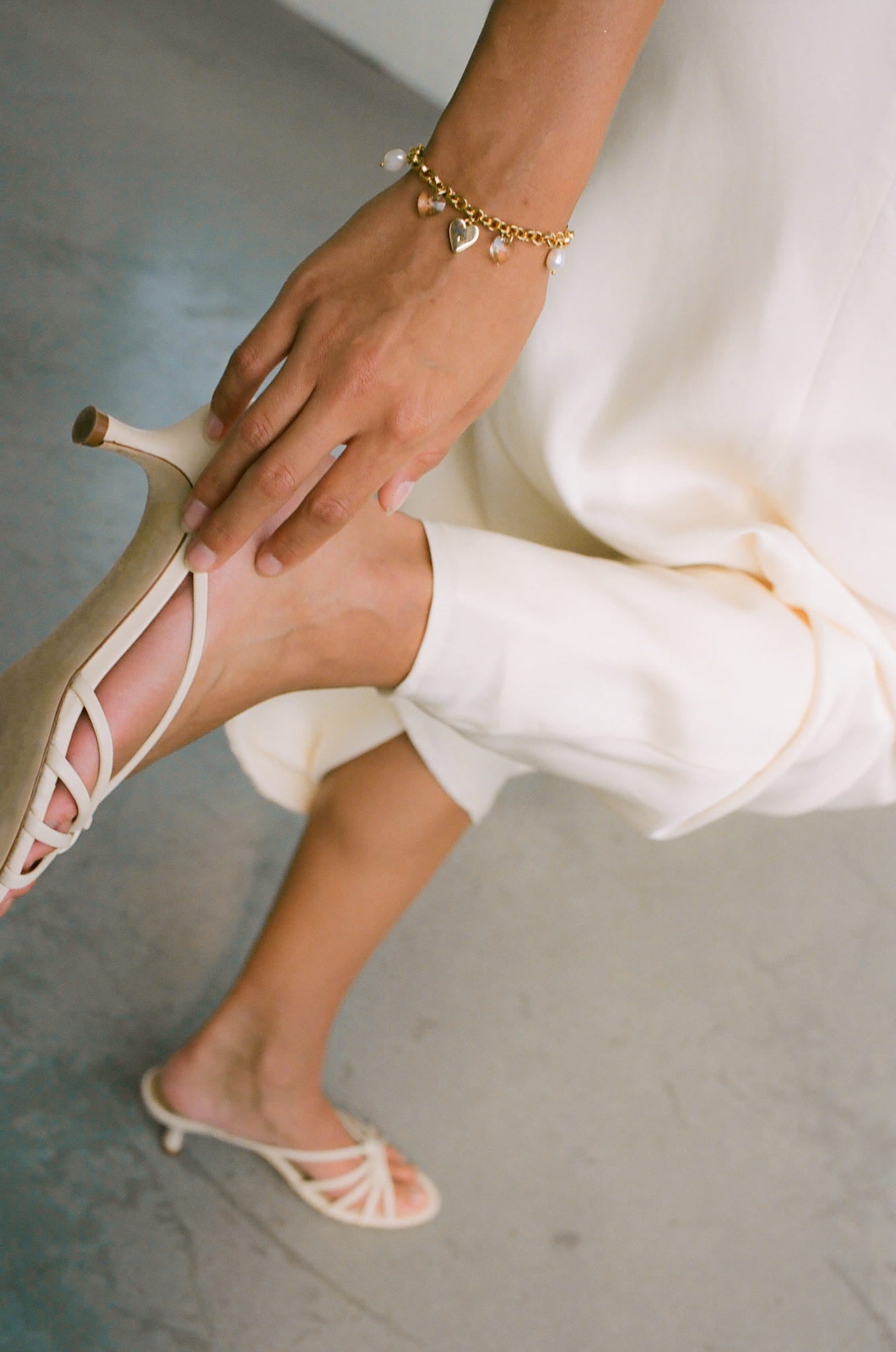 A person in cream-colored, strappy high heels and a matching loose-fitting outfit is holding their ankle, while their other foot is slightly lifted to showcase the Noémiah Secret Heart Bracelet. The bracelet features tarnish-resistant heart and pearl charms against a smooth, light-colored background.