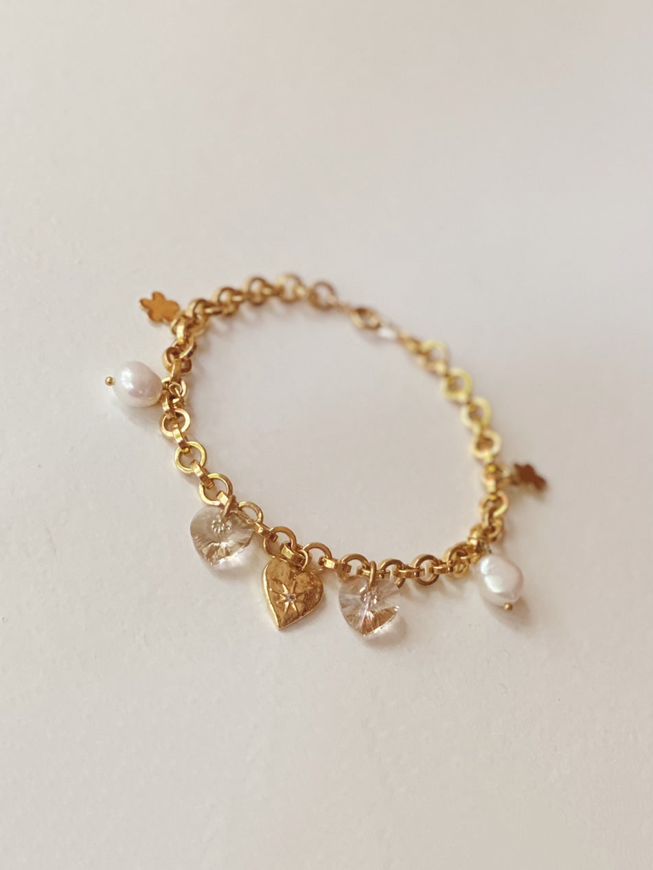 Introducing the Secret Heart Bracelet by Noémiah: a delicate gold accessory with interlocking links adorned by heart-shaped pendants, white pearls, and sparkling Swarovski crystals. Its tarnish-resistant finish guarantees a timeless elegant and refined look against a subtle light gray background.