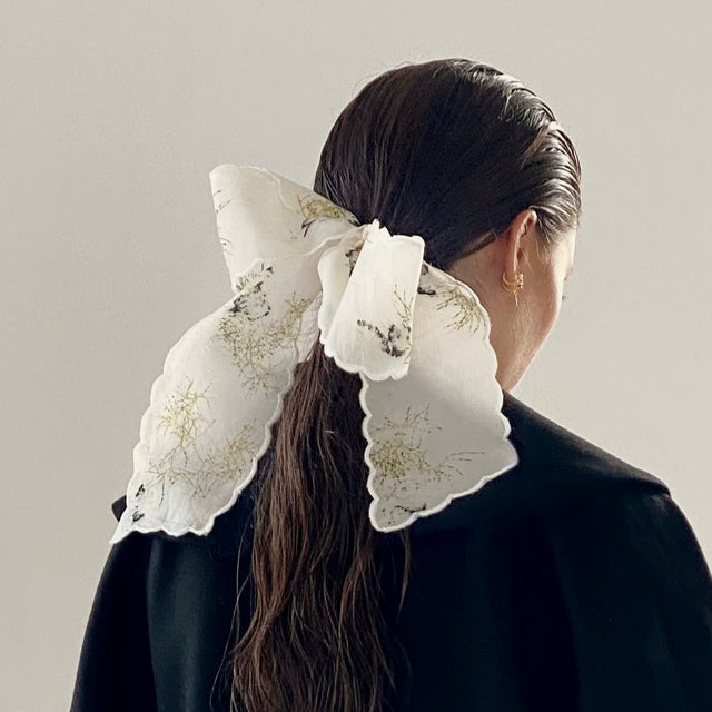 A person with long, dark hair is turned away, wearing a black coat and featuring the Noémiah Silk Barrette — Special Edition, adorned with floral patterns in their hair. The background is a plain light gray.