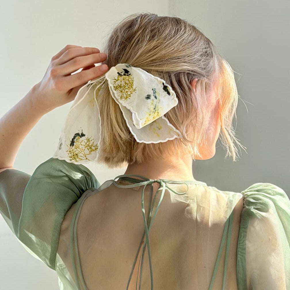 A person with short blonde hair, wearing a sheer green dress, faces away from the camera with their hair tied using Noémiah's special edition Silk Barrette, a handmade accessory embroidered with yellow and green floral details.
