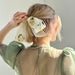 A person with short blonde hair, wearing a sheer green dress, faces away from the camera with their hair tied using Noémiah's special edition Silk Barrette, a handmade accessory embroidered with yellow and green floral details.