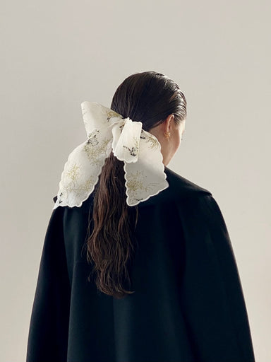 A person with long, dark hair is turned away, wearing a black coat and featuring the Noémiah Silk Barrette — Special Edition, adorned with floral patterns in their hair. The background is a plain light gray.