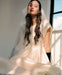 A woman with long dark hair wears a flowing cream-colored dress and black gloves. She sits indoors near a sunlit window, sheer silk organza draped around her shoulders. Her hair is adorned with the elegant Noémiah Silk Bow, enhancing the serene ambiance created by the soft lighting.