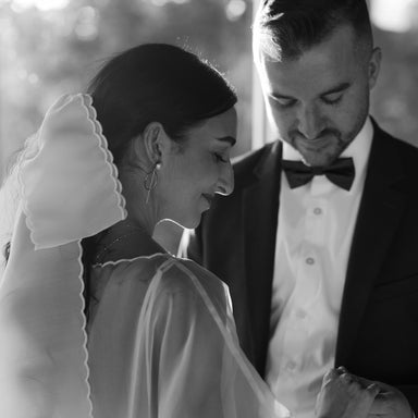 Bride in a sheer wedding dress with a Noémiah silk bow veil, holding hands with groom in a black tuxedo.