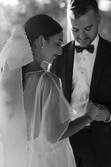 Bride in a sheer wedding dress with a Noémiah silk bow veil, holding hands with groom in a black tuxedo.