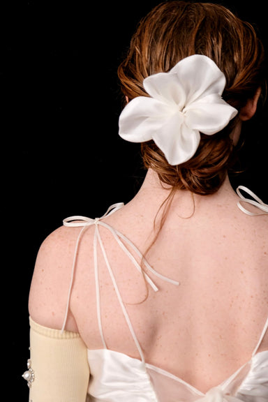 A woman with auburn hair styled in an updo, adorned with the Noémiah Silk Flower Comb, is shown from behind. She wears a white satin dress with delicate shoulder ties and a single cream-colored sleeve against a black background.