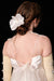 A woman with long hair styled in a low bun features a sculptural comb from Noémiah, embellished with a silk flower. She wears a white, silk dress accented by delicate tie straps and pairs it with long, cream-colored gloves adorned with pearls, set against a black background.