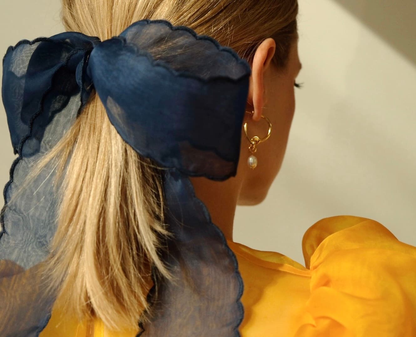 A person with blonde hair tied in a low ponytail, accented by an elegant navy blue silk ribbon from Noémiah. They wear a gold hoop earring adorned with a pearl and don a yellow blouse featuring puffy sleeves and embroidered edges. The background is a soft light shade, enhancing the sophisticated ensemble.