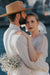 A couple embraces in a wedding setting. The groom, in a striped suit and brown hat, holds a bouquet of white flowers tied with a Silk Ribbon by Noémiah. The bride, in a white dress with embroidered edges and veil, gazes towards the camera. A blurred outdoor background is visible.