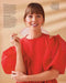 A woman models a stunning red silk dress from Noémiah's Silk Ribbon collection, featuring puffy sleeves and intricately embroidered edges, as she smiles at the camera. Her straight hair with bangs complements her large earrings beautifully. The background is a simple beige shade, accented by text on the left and in the top right corner of the image.