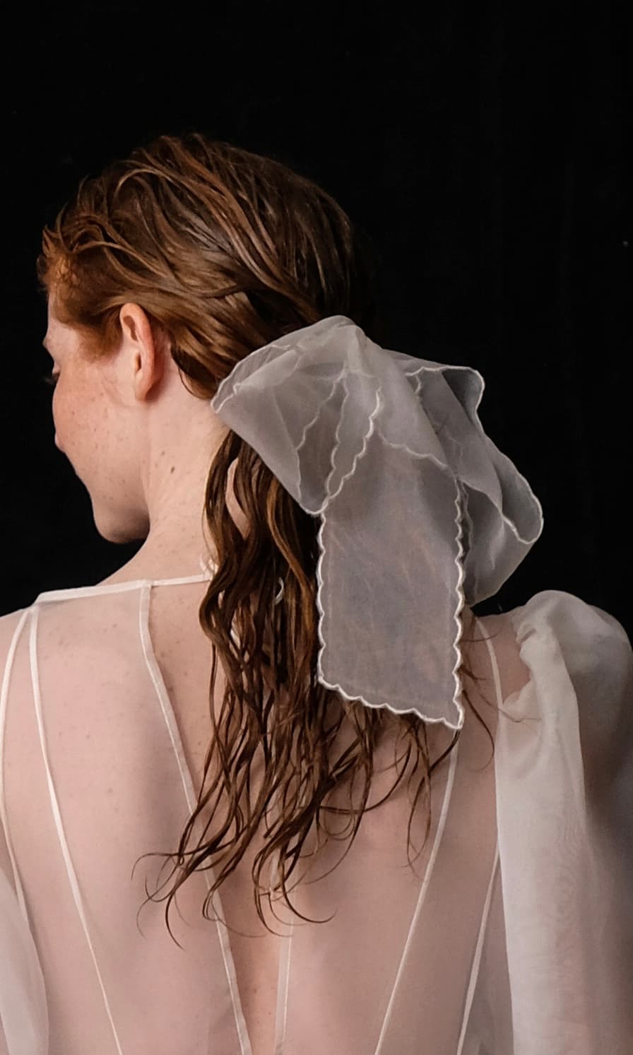 A woman with long brown hair is shown from the back, wearing a sheer white top with embroidered edges. Her hair is styled with an oversized Silk Ribbon by Noémiah. The background is black, adding contrast to the light colors of her outfit.