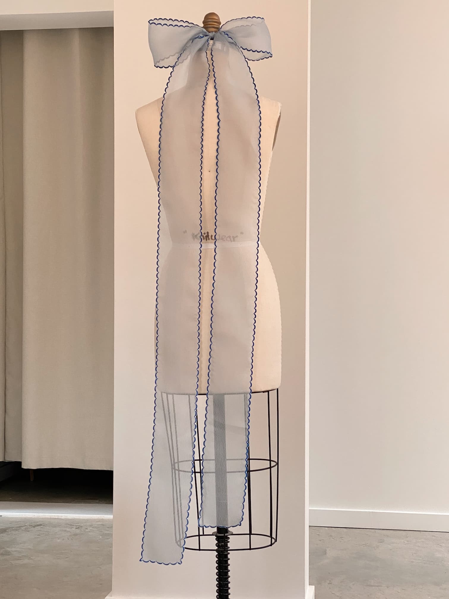 A dress form stands against a white wall, adorned with a Noémiah Silk Ribbon tied into a large bow at the top. The ribbon's long tails drape down the back of the mannequin, showcasing embroidered edges. The floor is concrete.